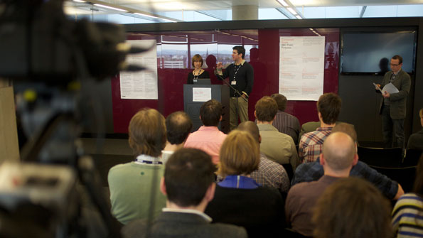 Two people speaking in a large open-plan meeting room. A video camera is out of focus in the foreground.