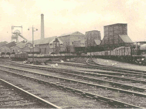 A steam train with a long line of wagons sits on one of a number of railway lines in front of industrial buildings including a winding tower and large chimney stack.