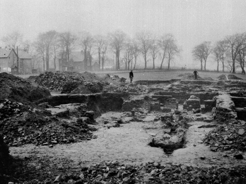 Black and white view of excavations of Roman fort at Mumrills. A line of trees and two large houses can be seen in the background.