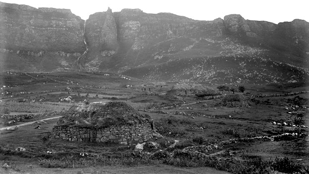 Black and white view of crofts with steep, rocky cliffs behind.