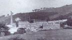 Black and white view of Atheenic Mill buildings and chimney stack, set on a largely grassy slope. There are two short terraces of houses by the mill buildings and a large detached house higher uphill.