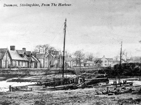 Black and white view of sailing boat in small harbour with row of cottages behind.