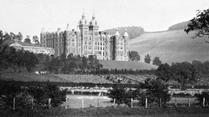 Black and white view of a large, five-storey hotel set on a wooded hillside above fields. The stone building is in Renaissance style, with a symmetrical frontage and a roofline featuring several turrets. A long conservatory extends from one side of the building.