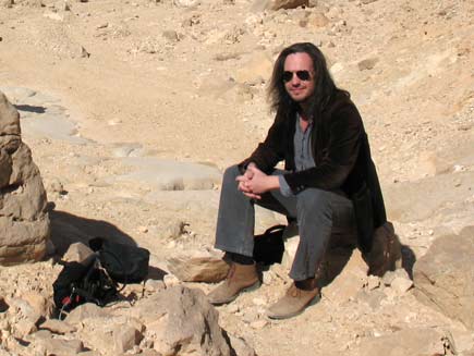 Nicholas Buxton, a long-haired man wearing sunglasses and dust-covered walking shoes, sits on a stone in the sun in the Egyptian desert