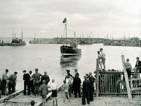 Black and white view of a boat being launched from a slipway into a harbour. A group of men and boys watch from the shore.