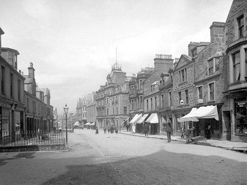 Black and white view of Crieff High Street, a wide, unsurfaced road lined by two and three-storey buildings with ground floor shops.
