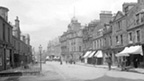 Black and white view of Crieff High Street, a wide, unsurfaced road lined by two and three-storey buildings with ground floor shops.