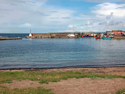 Colour view, taken on a sunny day, of a harbour which contains a number of small motor-powered fishing boats and pleasure craft. A single-masted fishing boat is tied up next to the empty quayside and slipway to the foreground.
