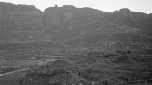 Black and white view of a natural rock amphitheatre in which lies a scattered collection of low, traditionally-built stone and thatch houses. Each house is surrounded by a low drystone wall.