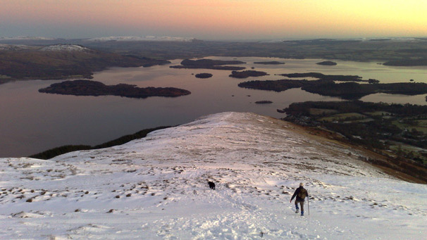 Hillwalker and dog descending in snow towards Loch Lomond