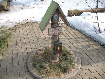 Memorial to Father Alexander: a small wooden pillar set in the middle of a paved path, surrounded by snow. The display has a tiny roof to protect an image of Jesus and a plaque bearing Alexander's name from the elements. A lighted lantern is hanging underneath the plaque