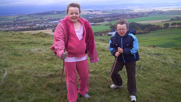 Two children battle the wind to stay standing on a hilltop