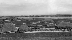 Black and white view of low, stone cottages with thatched roofs along the sides of a small loch. A larger, two-storey, pitched roofed building with slate or tile roof can be seen in the background.
