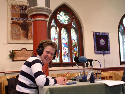 Clair Jaquiss smiles from her place at a tiny desk, on which are several microphones, scripts, pens and a clock