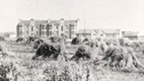 Small haystacks in a wide field with a tenement block and other buildings beyond.