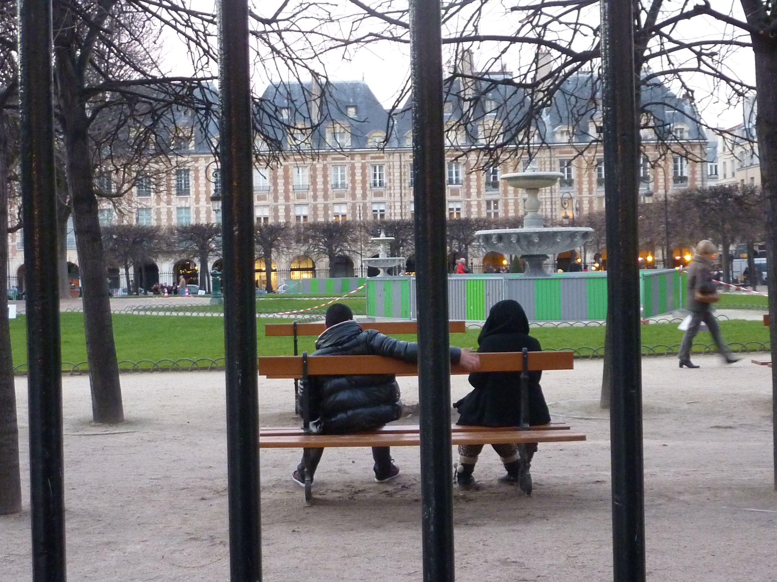A young couple on a bench in a park. The young man extends his arm towards the girl. 