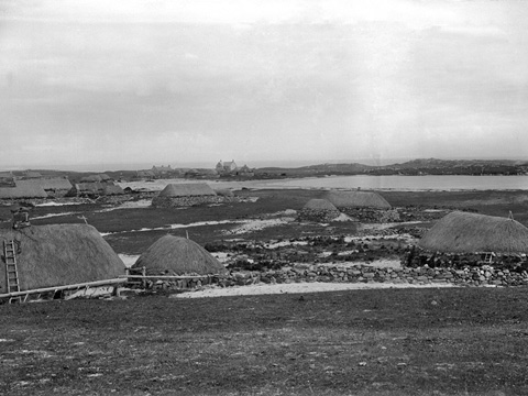 Black and white view of low, stone cottages with thatched roofs along the sides of a small loch. A larger, two-storey, pitched roofed building with slate or tile roof can be seen in the background.