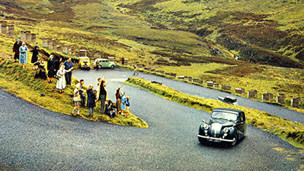 Colour image of a vintage Daimler motorcar climbing a steep, double-hairpin section of road in a mountainous area of grass and heather. A small crowd of people stand waving at the roadside.