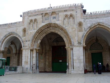 Facade of the Al-Aqsa mosque, a stone building on square lines with three arched doorways. Visitors stand around talking and security lights are visible on the corners of the roof