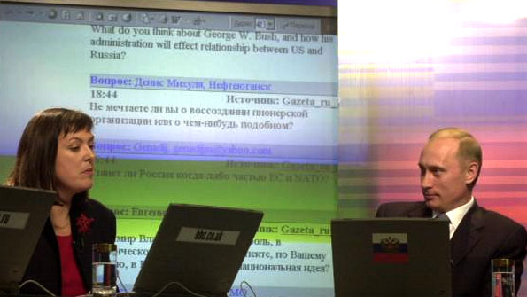 A female reporter and Vladimir Putin in a brightly coloured studio. Laptops in the foreground, and scrolling chat text - in Russian and English - behind them.