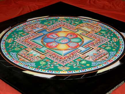 A view of the mandala, now close to completion. The pattern contains squares and circles and is symmetrical in four directions. The dominant colour is green, with red, blue and yellow sand and many details in black and white. The middle section represents a building and green areas nearer the edge resemble gardens filled with trees and other designs