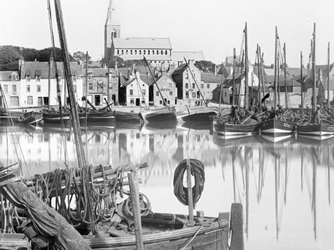 Black and white view of harbour filled with sailed fishing boats with town dominated by church tower in the background