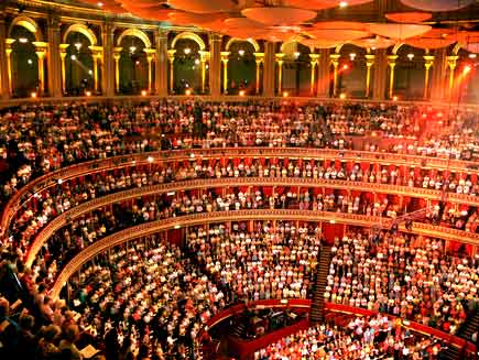 Audience members are seated in three gallery levels and on the main level of the circular Royal Albert Hall, illuminated by spotlights from the arches around the top edge