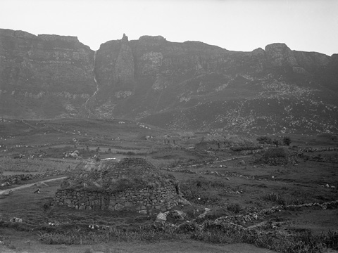 Black and white view of a natural rock amphitheatre in which lies a scattered collection of low, traditionally-built stone and thatch houses. Each house is surrounded by a low drystone wall.