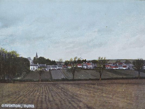 Hand-tinted view across gently rolling, ploughed fields to a village beyond. A church spire rises to the left of frame.