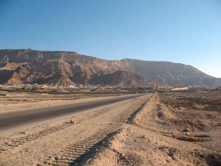 A road through the Egyptian desert. Tyre marks can be seen in the sand on the tarmac. In the distance a collection of low, light-coloured stone buildings is dwarfed by the red-brown mountains behind