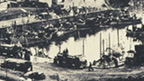 Black and white view of harbour with fleet of fishing boats and a number of loaded trucks on the quayside.