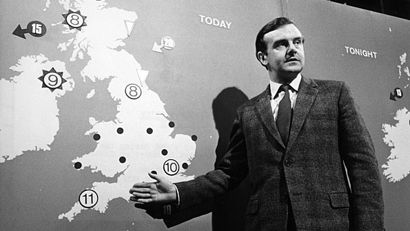 B/W: Man in suit stands in front of map of Britain.