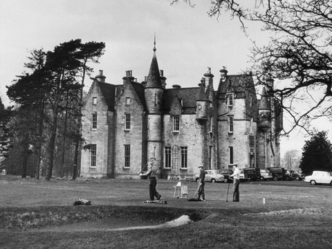 Black and white view of three men playing golf, with a large Scottish Baronial house and trees behind.