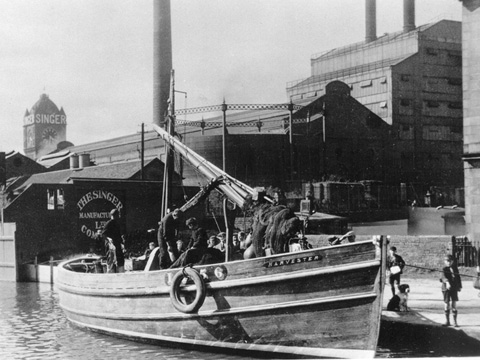 Black and white view of extensive industrial buildings with tall smoking chimney stacks with tall, flat topped hills behind.