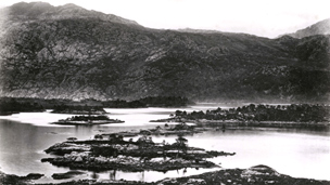 Black and white view looking across the islands of Loch Maree to mountains beyond.