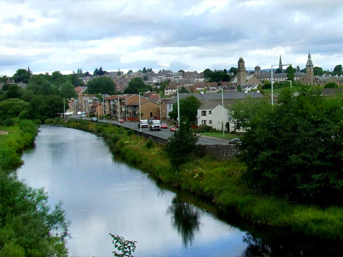 Colour view of wide flat river with a town to the right of frame. A mix of modern houses and low flats line a road next to the river. The bank to the left of frame has trees, grass and a footpath.