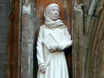 Statue of Julian of Norwich, a small, fine-boned woman in mediaeval kirtle and headscarf, holding a quill pen and a book with the title Revelations of Divine Love, with the wall and arched door of a cathedral visible in the background