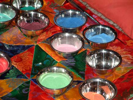 Polished bowls full of coloured sand are arranged on a colourful cloth by the edge of the mandala table