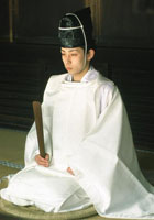 Shinto priest in white robes and black ceremonial hat, kneeling on a straw mat and holding a wooden wand in his right hand