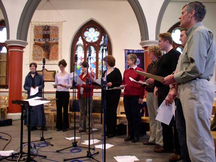 The Daily Service Singers, nine men and women wearing ordinary clothes, stand in a semicircle around small microphone stands inside the church. Christian-themed wall hangings and stained glass windows can be seen in the background