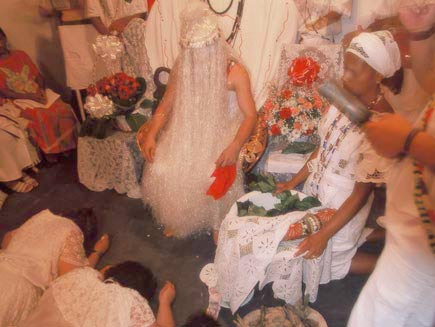 Man sitting on chair with his head covered by silver fringe all the way to the floor. Female worshippers prostrate before him.