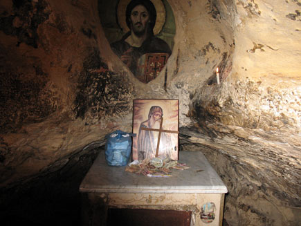 Inside the cave a small table serves as altar, with a plastic bag of papers, a pile of monetary donations, a painting of Saint Antony and a simple wooden cross. On the wall behind, seeming to be painted directly on the stone, is a larger picture of Christ. A small white candle burns in a niche in the rock