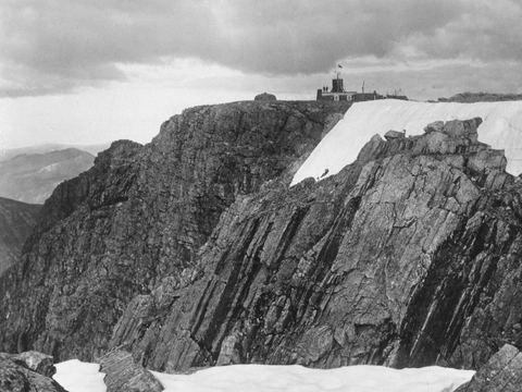 Black and white view across rocks, snow and ice to the flat summit of Ben Nevis. At the summit stands a small, low building with a central tower.
