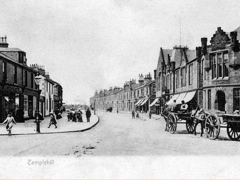 Black and white view of a couple with a pram and dog walking along a winding, coastal footpath. In the background are trees and the tall, cylindrical Battle of Largs Monument.