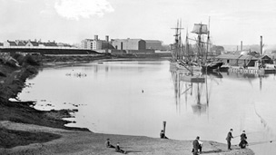 Black and white view of Perth Harbour from the south. A tall sailing ship is docked at the harbour which protrudes in to the river. In the foreground, a group of men are fishing while another man in a top hat sits sketching or painting the scene. In the background are some large, possibly industrial buildings.