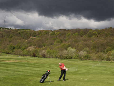A single golfer stands mid swing on a sunny golf course with tall, flat topped hills behind under a threatening sky of dark clouds.