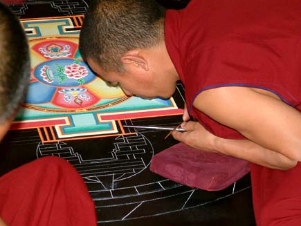 Monks, dressed in red tunics with close-cropped hair, lean over a circular design marked out in chalk on a black tabletop. Their faces are inches from the surface. Resting their hands on small cushions, they are pouring coloured sand through small metal tubes into the spaces marked by the chalk outline