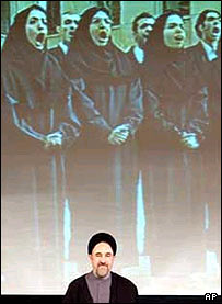 محمد خاتمی ايستاده در کنار پرده تلويزيونی بزرگی، به سرود رسمی ايران که توسط گروهی از دانش آموزان اجرا می شود گوش فرا می دهد