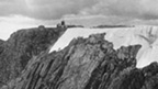 Black and white view across rocks, snow and ice to the flat summit of Ben Nevis. At the summit stands a small, low building with a central tower.