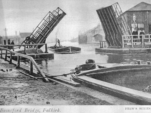 Black and white view from canal side showing a barge passing under an open bascule bridge.